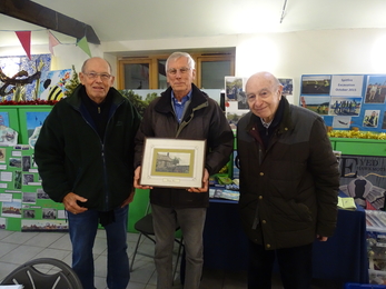 Three gentlemen stand with the centre man holding a framed photograph 