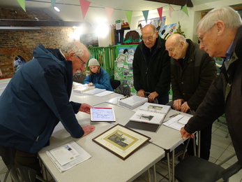 4 men stand around tables looking at a map on a tablet and folders of photographs