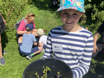 Evie stands smiling to camera behind a black bucket containing pond plants, rocks and water