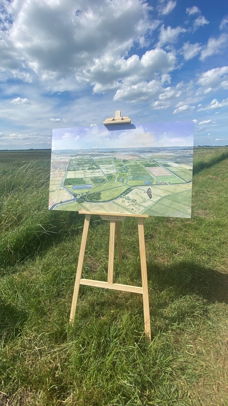 A painting of green landscape and blue skies is on a wooden easel standing on a grass bank with fields behind. 