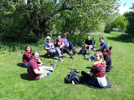 A group of adults sit in the sun enjoying their packed lunches