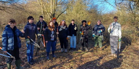 A group of 10 young people holding tool such as loppers and saws stand in woodland smiling to camera