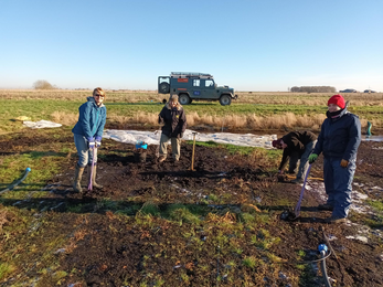 4 adulst stand on muddy field with spades, blue sky and a land rover parked in the distance