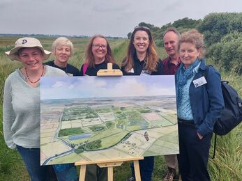 5 ladies and 1 man stand smiling to camera on the bank at Woodwalton Fen behind the Peatland Progress artwork on an easel