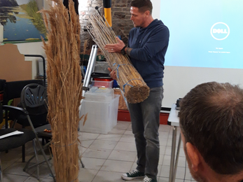 A man stands inside a classroom holding a bundle of reed next to another bundle on the floor