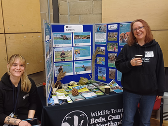 Rebekah stands and Lara sits next to a table and display board with leaflets and pictures about the Wildlife Trust