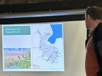 A man stands in front of a slide on a projector screen showing the location of RSPB Ouse Fen on a map