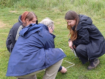 Two young woman with CEO Brain Eversham kneeling in a field, looking into a quadrat