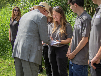A line of young people with a Wildlife Trust work clothes. One girl is holding out a book that the Duke of Gloucester is signing.