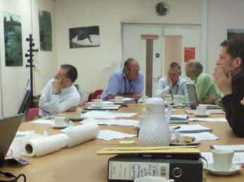 A group of men sit around a large table discussing paperwork and one man stands pointing at a map of the Great Fen