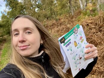Ellen takes a selfie standing in a birch woodland holding a fungi ID guide, 