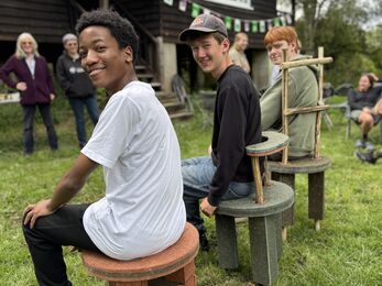 Three teen boys sit on stools they have made. They look over their shoulder to camera. 