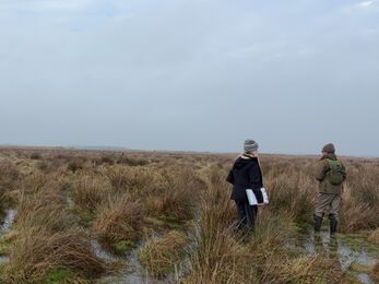 A young woman and a man in wellies standing in a flooded field holding a pair of binoculars and a clipboard