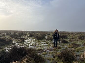 A young woman in wellies standing in a flooded field holding a pair of binoculars smiling