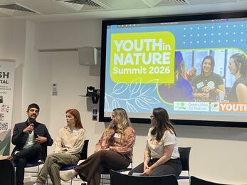 A man and three women sit in a row speaking to an audience in front of a screen showing the Youth in Nature Summit logo