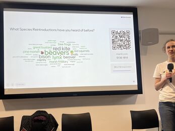 A speaker with microphone presents a slide on a large screen asking which species reintroductions people had heard of