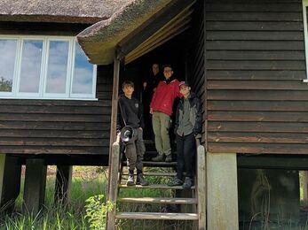 Three young men and a woman stand on the steps to a wooden bungalow on concrete stilts