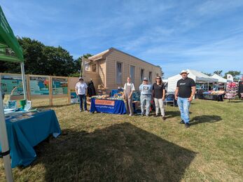 Five staff members stand in front of the wooden tiny house on a sunny day in a field at a trade show