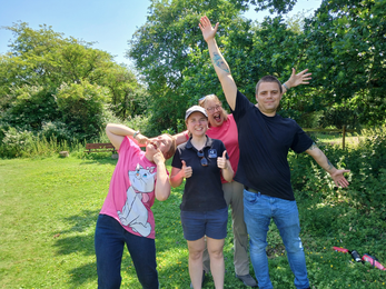 Laura, Tansy, Lewis and their support worker in the sunshine at the reserve. Tansy pulls a funny face, Lewis and the support worker stretch their arms wide, Laura gives a double thumbs up and 