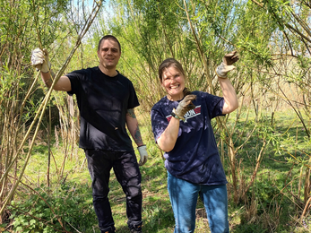 Tansy and Lews, wearing tshirts and work gloves, stand smiling inside a willow tunnel on a sunny day
