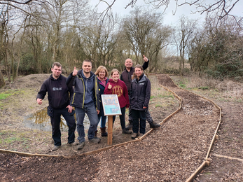 Three men and two women smile to camera in a nature reserve beside a new woodchip path, they give thumbs up
