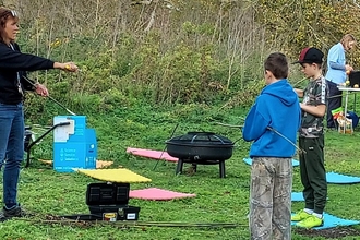 Lady standing facing two boys, showing them how to hold and use a knife safely for whittling