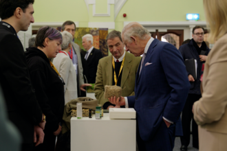 King Charles holding a block of sphagnum moss in one hand, other hand in his pocket. He speaks with Lorna Parker, standing opposite. Other people stand around them in a large hall.  