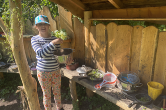 Girl holding pan and plants by wooden mud kitchen