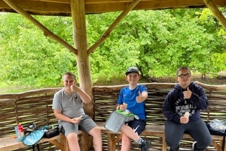 Three boys sit in a wooden shelter giving a thumbs up to camera