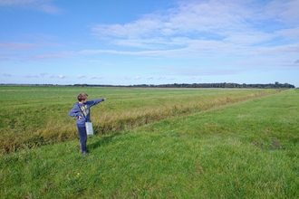 A boy in a blue jumper stands in a green field pointing to a tree line in the distance