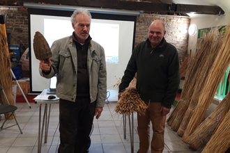 Two men stand inside the classroom at Ramsey Heights, one holding a bundle of reed and one holding an old tool for thatching work