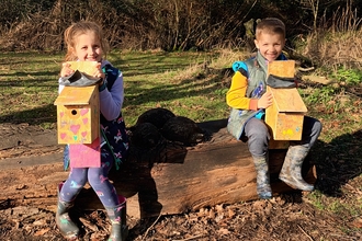A young girl and boy sit on a wooden log bench holding the bird boxes they've made. 