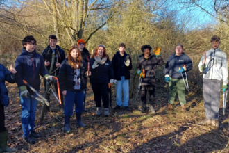 A group of 10 young people holding tool such as loppers and saws stand in woodland smiling to camera