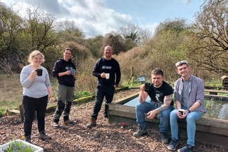 Three people stand and two sit on the edge of a raised pond, all have mugs of tea. It is a sunny day. 