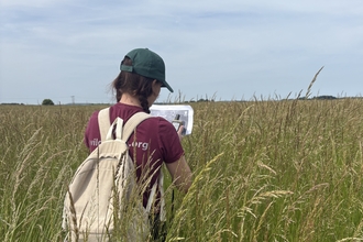Lucy walks through a shoulder height field of grass holding a GPS tracker and map