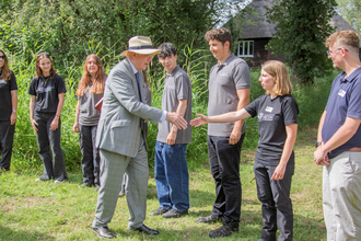 A line of young people with a Wildlife Trust work clothes. The Duke of Gloucester is reaching out to shake a hand of a young woman