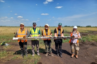 Five men in high vis waistcoats hold a long soil core, they stand in a flat fenland field, blue sky