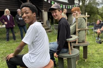 Three teen boys sit on stools they have made. They look over their shoulder to camera. 