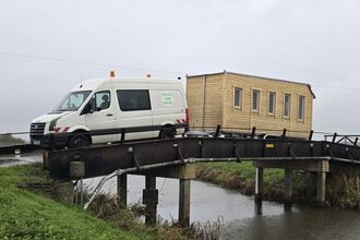 A wooden tiny-house is pulled over a bridge by a white van
