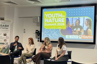 A man and three women sit in a row speaking to an audience in front of a screen showing the Youth in Nature Summit logo