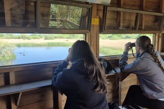 Two teen girls sit in a bird hide using binoculars, a parabolic microphone on the counter