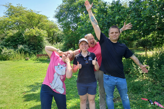 Laura, Tansy, Lewis and their support worker in the sunshine at the reserve. Tansy pulls a funny face, Lewis and the support worker stretch their arms wide, Laura gives a double thumbs up and 