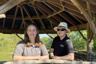 Aimee and Jake stand inside a wooden domes structure outside in a nature reserve. They smile to camera and lean on a half-height wall panel made of woven hazel and willow. Aimee wears a grey Wildlife Trust tshirt and Jake's is black. Jake also has sunglasses and a cream panama hat. 