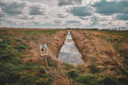 View over flat green and yellow grassland peatland with trees in distance, water filled drain centre and a deep water warning sign in the foreground