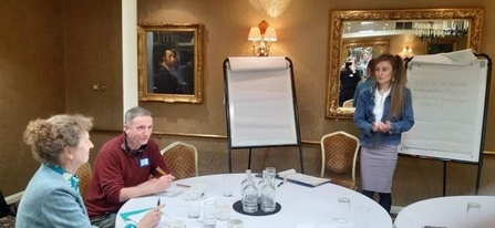 Adult female and male sat at round table looking at adult female standing by two flip charts