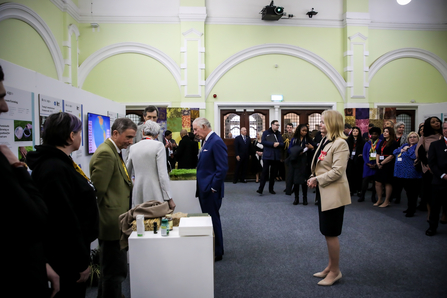 King Charles looking at exhibits in the Great Hall. The hall has yellow walls with white arches. The King wears a blue suit. Lots of people dressed smartly stand around. 