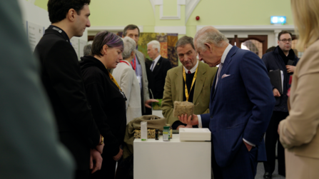 King Charles holding a block of sphagnum moss in one hand, other hand in his pocket. He speaks with Lorna Parker, standing opposite. Other people stand around them in a large hall.  