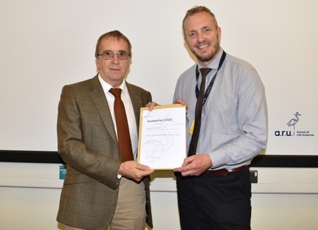 Two men stand holding a certificate between them, smiling to camera