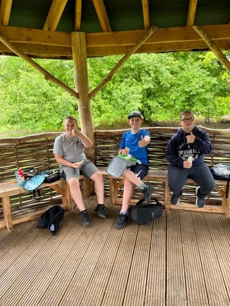 Three boys sit in a wooden shelter giving a thumbs up to camera