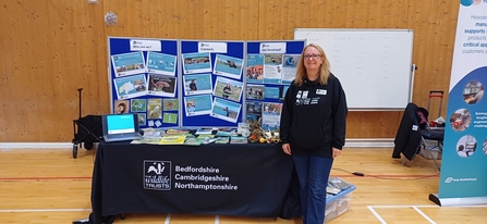 Rebecca stands wearing a Wildlife Trust black hoodie in front of a table and display board showing event posters, leaflets and a laptop